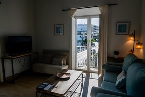 Una sala de estar con un sofá, una mesa de centro con un cuenco y un control remoto, un televisor sobre un mueble bajo, y un balcón con barandilla.