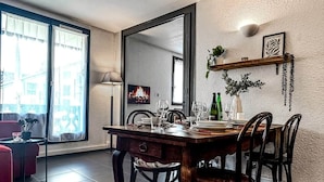 Uma área de jantar com mesa de madeira posta para quatro, cadeiras, um sofá vermelho, uma mesa pequena com uma planta e um espelho grande na parede.