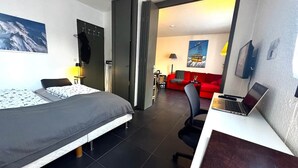Quarto de hotel com cama, escrivaninha, laptop e um sofá vermelho ao fundo.