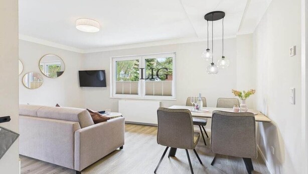 Een moderne woonkamer met een bank, een eettafel met stoelen, een flatscreen-televisie aan de muur en een raam met zonwering.