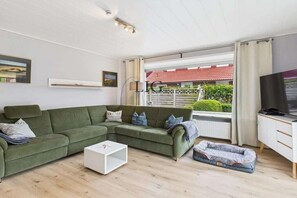 A modern living room with a sectional sofa, a white coffee table, a pet bed, a television, and a window with a view of a red-roofed building. - Holiday apartment Strandkoje (Langeoog)
