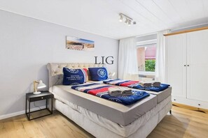 A bedroom with a bed, a nightstand, a lamp, a window with curtains, and a door. - Holiday apartment Strandkoje (Langeoog)
