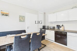 A modern kitchen with white cabinets, a dishwasher, and a microwave. There is a dining table with chairs and a clock on the wall. - Holiday apartment Strandkoje (Langeoog)