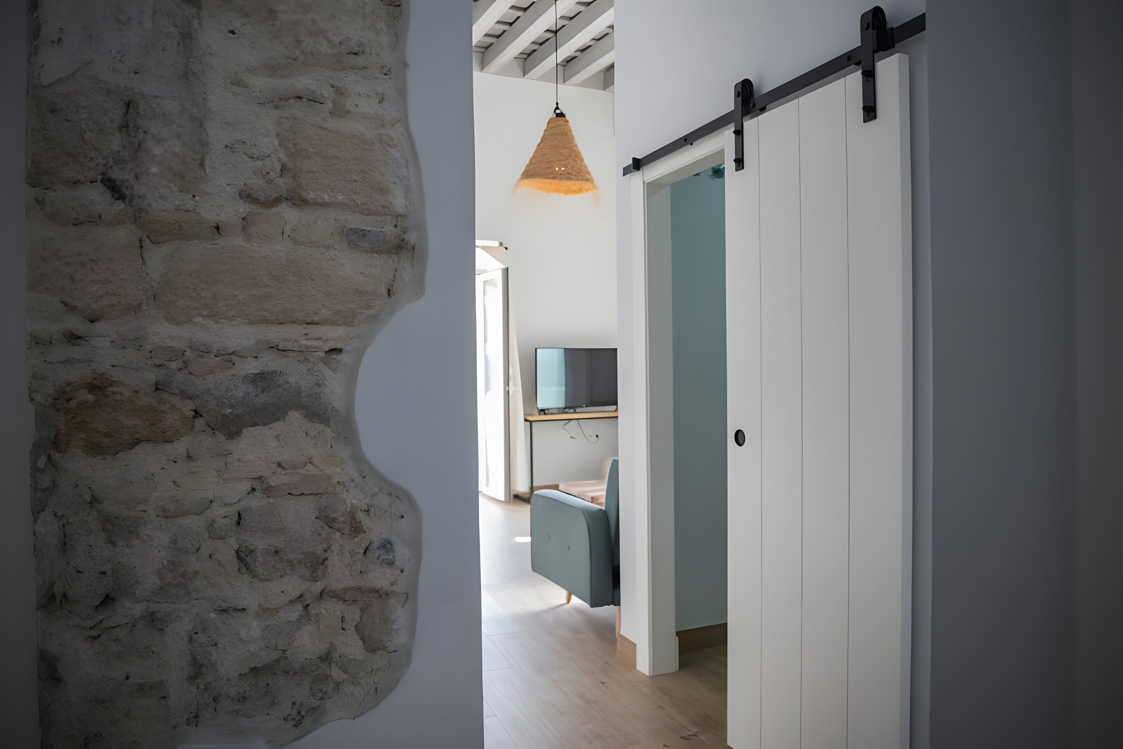 Un couloir avec un mur en pierre, une porte coulissante de style grange, une suspension lumineuse et un aperçu d’une pièce contenant un bureau et une chaise.