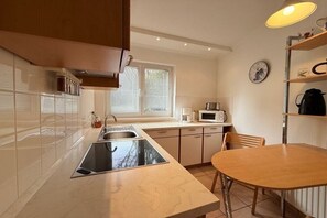 A modern kitchen with a sink, stove, and microwave. There is a dining table with chairs and a window with blinds. - Holiday home with 1 bedroom (Travemünde)