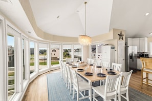 A dining area with a large table set for six, surrounded by white chairs, with a view of the outside through large windows. - Elevator, Theater Room, Game Room, Duck, Oceanfront, Pool, D0128 (Duck)