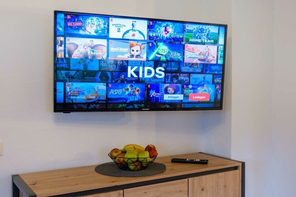 A flat-screen TV mounted on a wall displaying various children's shows and games, with a fruit bowl and remote control on a wooden cabinet below.