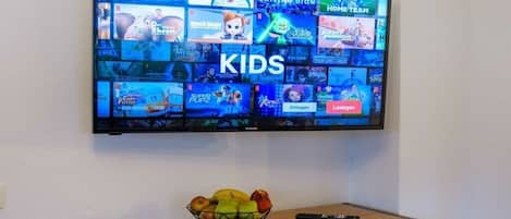 A flat-screen TV mounted on a wall displaying various children's shows and games, with a fruit bowl and remote control on a wooden cabinet below.