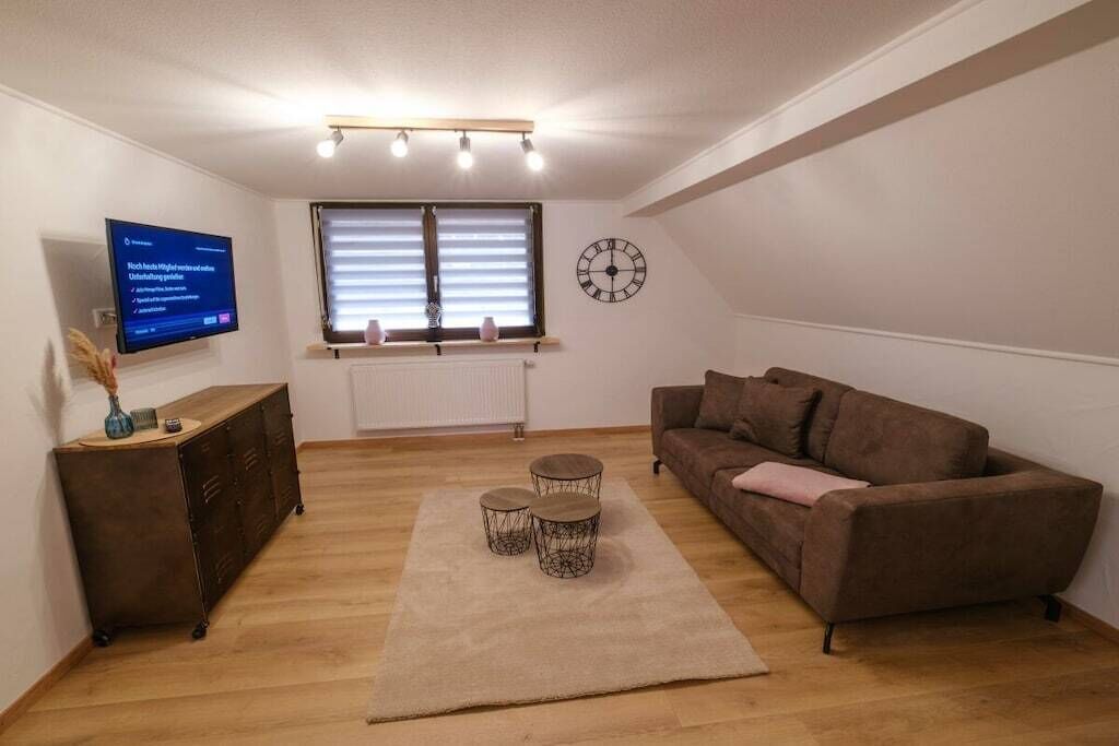 Un salon avec un canapé marron, un téléviseur fixé au mur, une fenêtre avec des stores et une horloge.