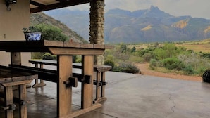 An outdoor patio with a stone pillar, wooden table, and benches, overlooking a mountainous landscape.