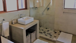 Casa de banho com chuveiro de cabine de vidro, lavatório branco e janela sobre o espelho.