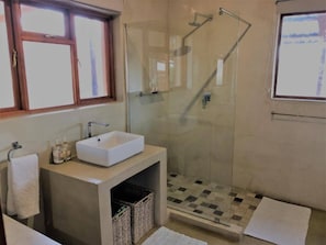 Een badkamer met een douchecabine van glas, een wit wastafeltje en een raam boven de spiegel.