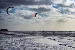 A beach with a structure in the distance, a person kite surfing, and another person swimming. - Holiday home with 3 bedrooms (St.Peter-Ording)