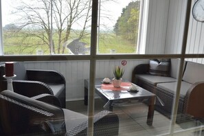Ein Zimmer mit Korbmöbeln, einem Glastisch und Blick auf ein Feld durch ein Fenster.