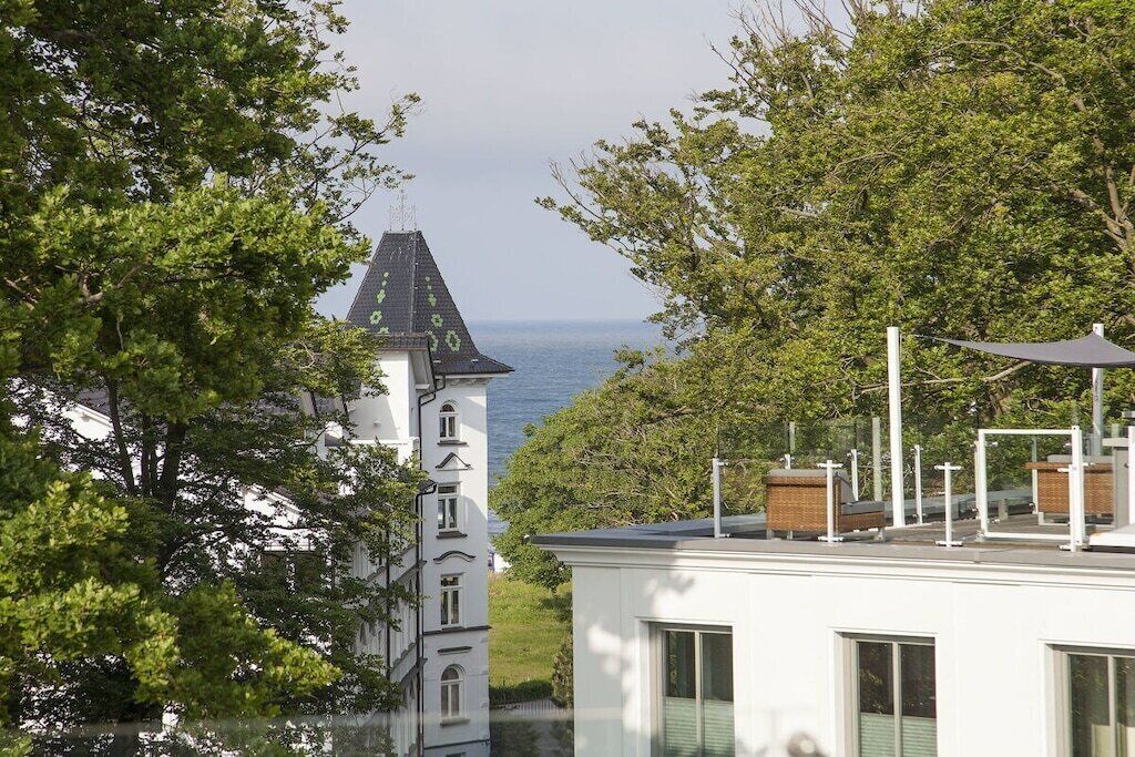 A white building with a tower, a view of the sea, and trees in the foreground.