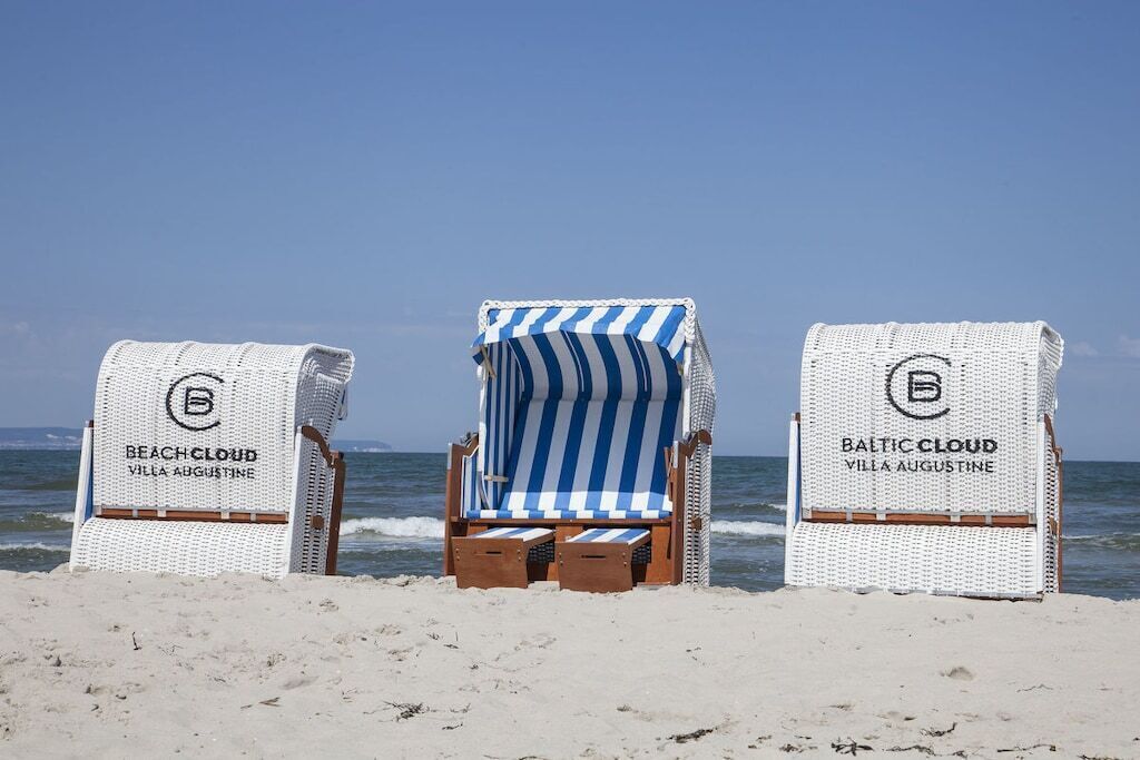 Three beach chairs with towels, labeled 'BeachCloud Villa Augustaine' and 'BalticCloud Villa Augustaine,' on a sandy beach with the ocean in the background.