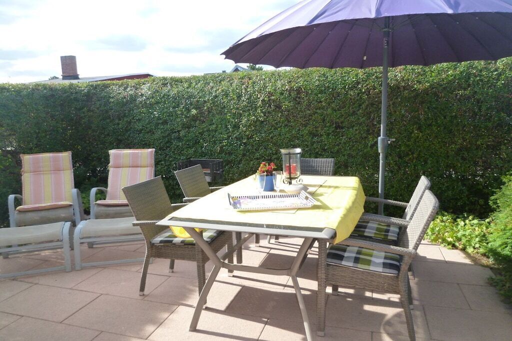 An outdoor patio with a table and chairs, a large umbrella, and a hedge in the background.
