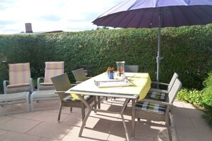 Een terras buiten met een tafel en stoelen, een grote parasol en een haag op de achtergrond.