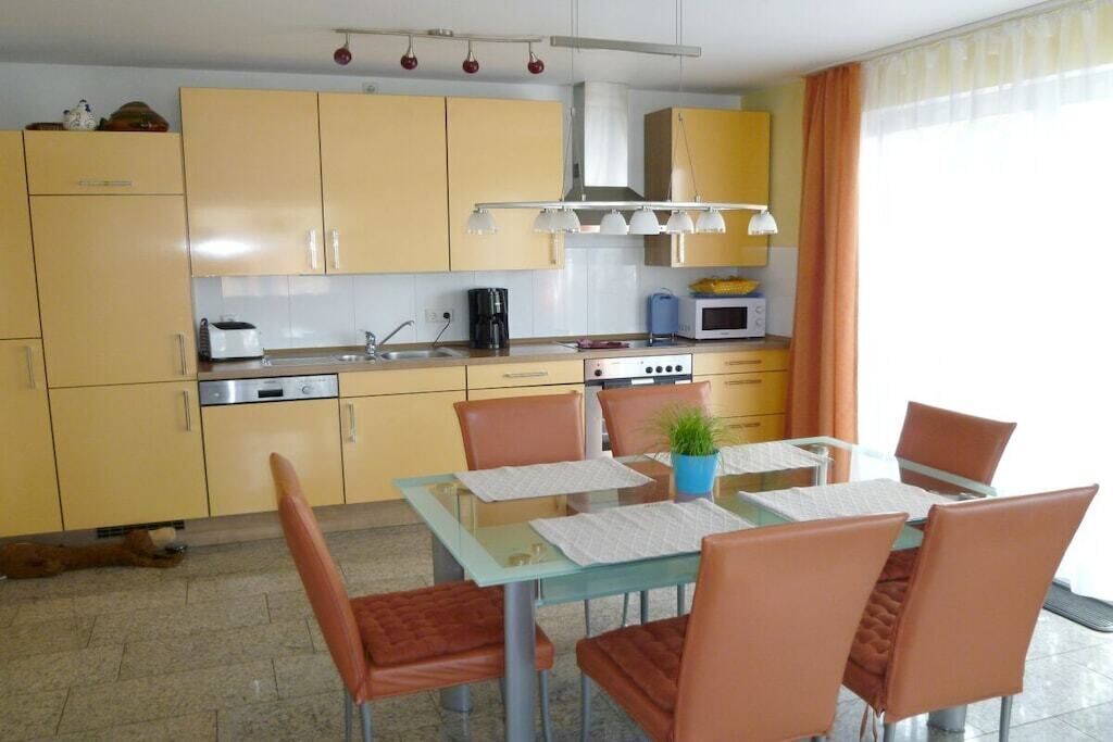 Uma cozinha compacta com armários amarelos, uma mesa de jantar com cadeiras e uma pequena janela com cortinas.