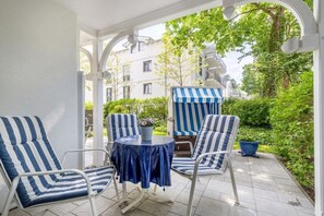 A covered patio area with striped lounge chairs and a small round table, surrounded by greenery and a building in the background. - 2 in the Granitz house (Binz)