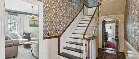 A staircase with a wooden handrail and black steps, leading to a room with patterned wallpaper and a chandelier.