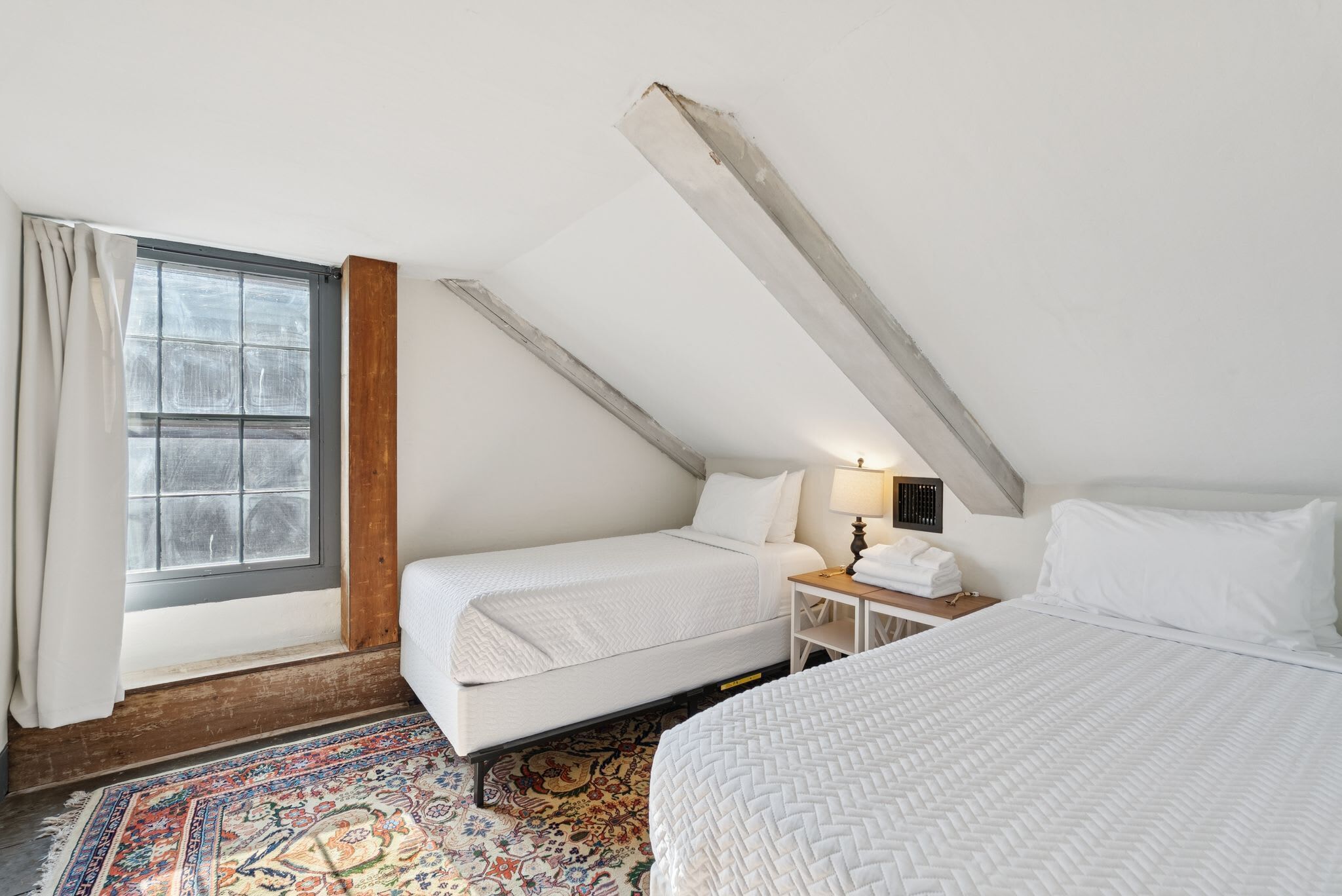 A room with a sloped ceiling, a bed, a nightstand, a lamp, and a window with curtains.