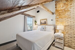Una camera da letto con soffitto spiovente, un letto con biancheria bianca, un comodino con una lampada e un muro in mattoni.
