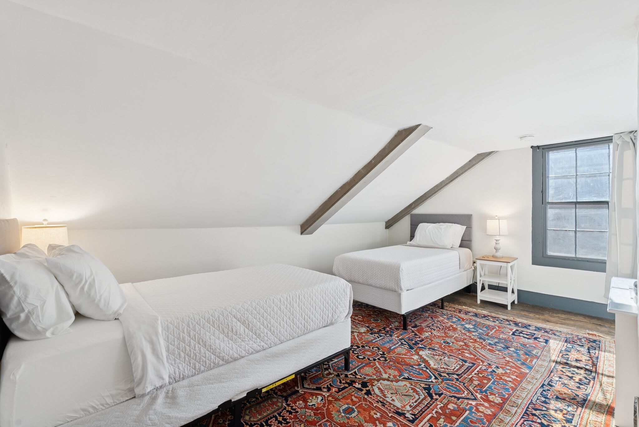 Een slaapkamer met een groot bed, een kleiner bed, een nachtkastje, een lamp, een raam en een kamerbreed tapijt met dessin.