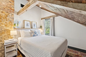 A bedroom with a brick wall, a wooden ceiling beam, a bed with white bedding, a nightstand with a lamp, and a window with curtains.