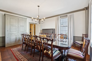 A dining room with a glass table surrounded by wooden chairs with striped cushions, a chandelier, and a large window with curtains.