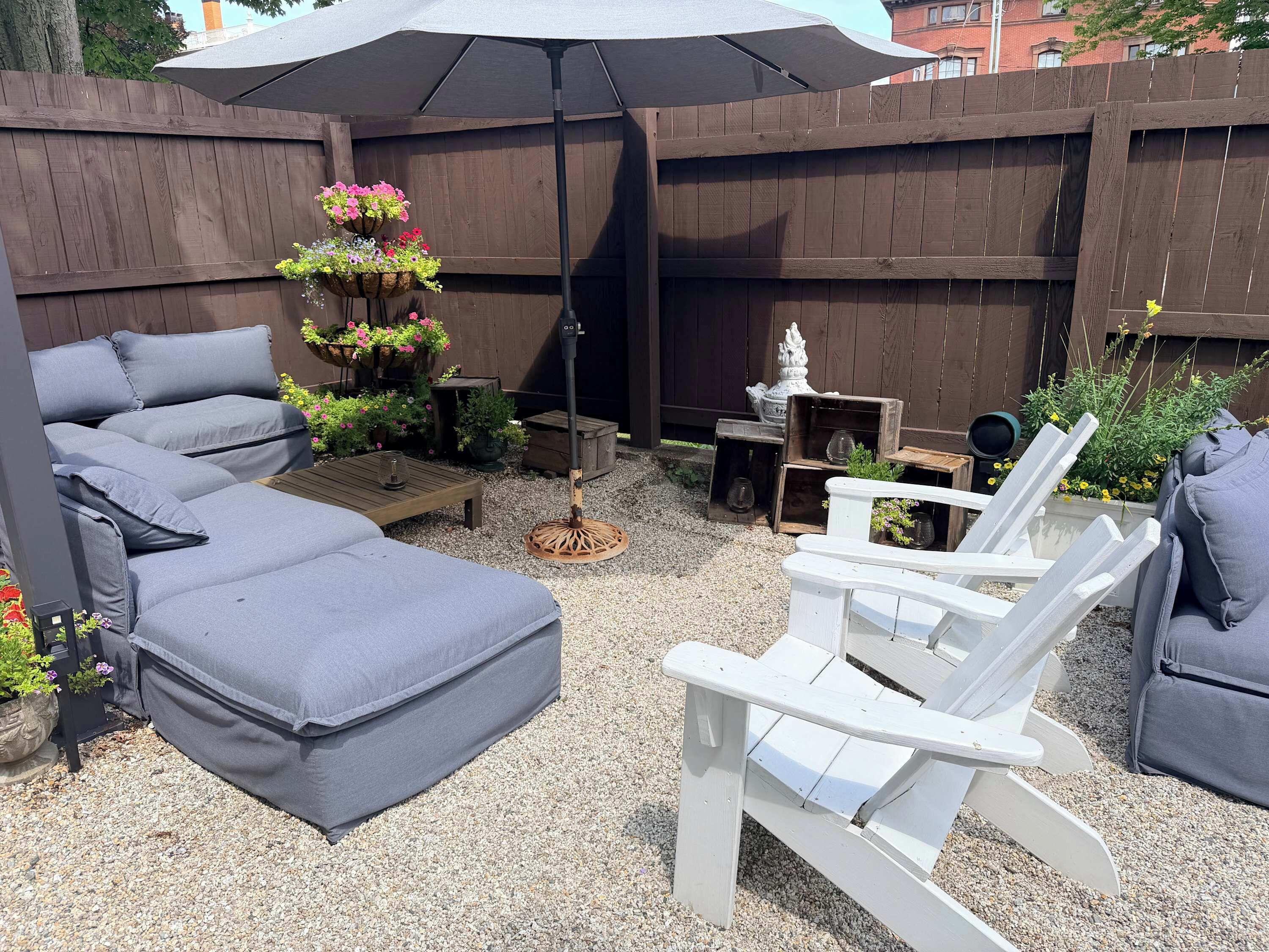 Een achtertuin met een grijze bank, witte Adirondack-stoelen, een houten tafel en een hangende bloemenmand.