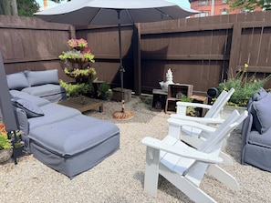 A backyard with a grey sofa, white Adirondack chairs, a wooden table, and a hanging flower basket.