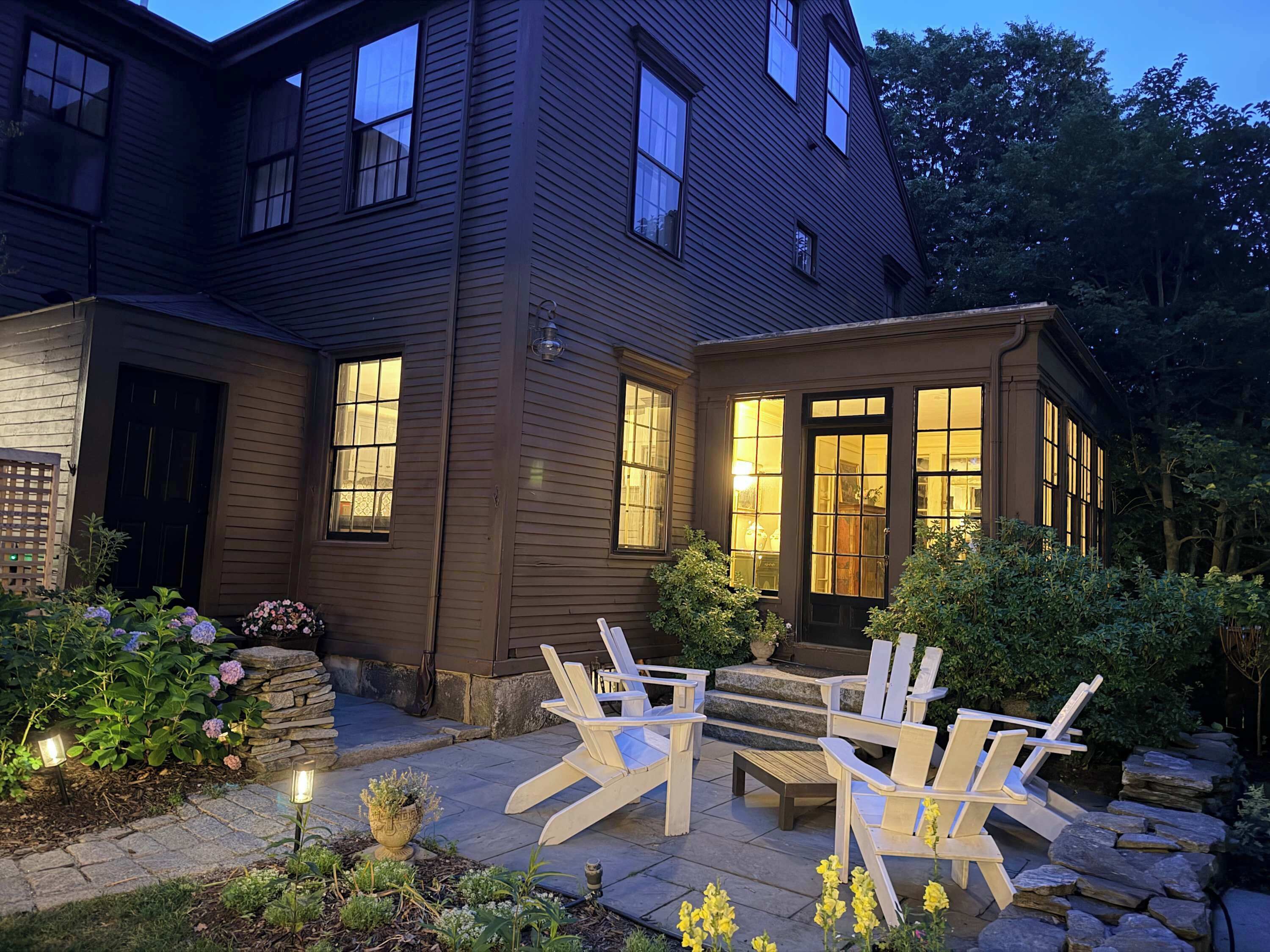 A two-story house with a stone staircase leading to a front door, surrounded by a garden with potted plants and a seating area with chairs.