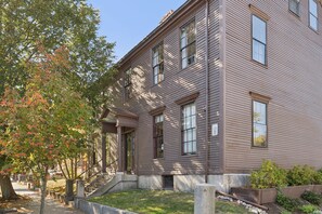 A two-story building with a traditional architectural style, featuring a front entrance with a set of stairs leading up to it, and a small front yard with a tree and a planter. - The Diamond Island Suite - Suite in Historic Home (Portland)