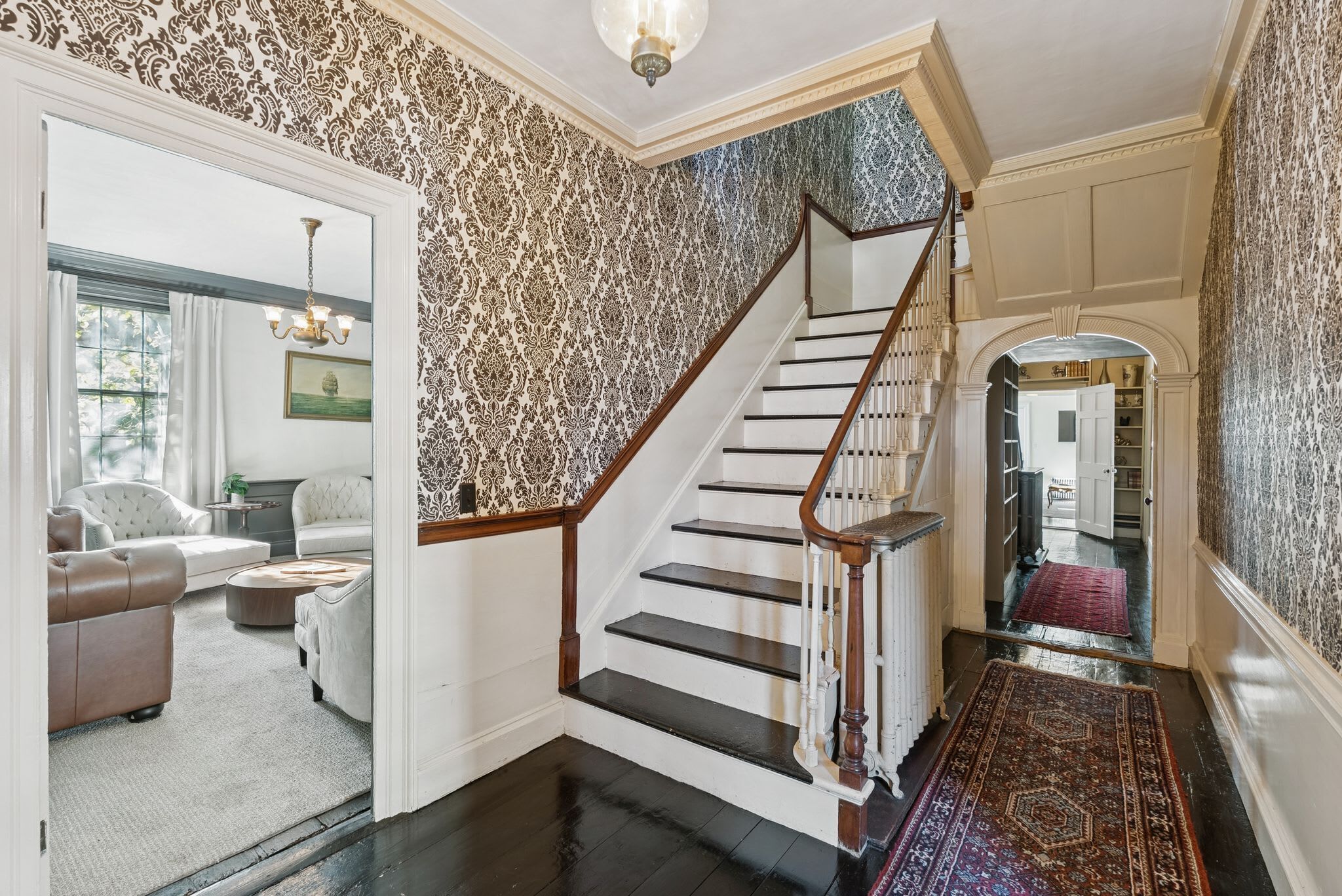 A staircase with a wooden handrail and black steps, leading to a room with patterned wallpaper and a chandelier.