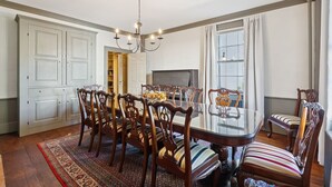 A dining room with a glass table surrounded by wooden chairs with striped cushions, a chandelier, and a large window with curtains.