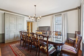 A dining room with a glass table surrounded by wooden chairs with striped cushions, a chandelier, and a large window with curtains.