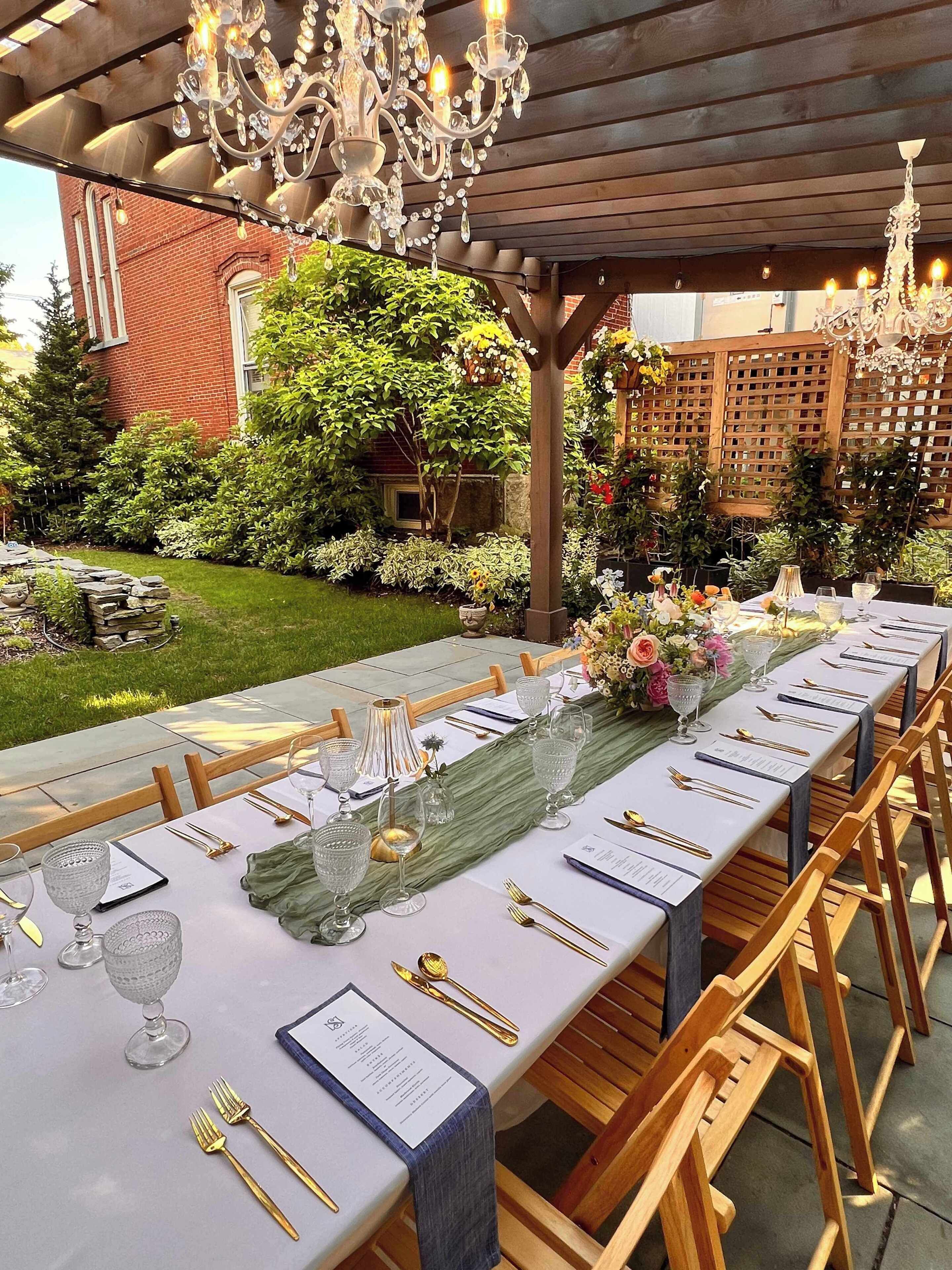 Een buitenterras met een lange tafel gedekt voor een maaltijd, omgeven door groen en een houten pergola.
