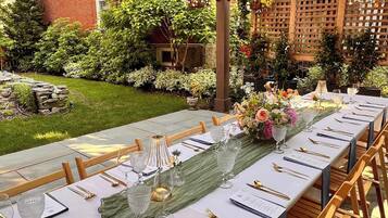 Een buitenterras met een lange tafel gedekt voor een maaltijd, omgeven door groen en een houten pergola.