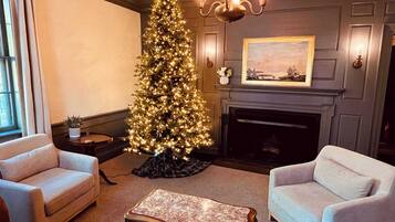 Een gezellige woonkamer met een hoge, verlichte kerstboom, twee fauteuils, een marmeren salontafel en een open haard met daarboven een lijstwerk met een schilderij.