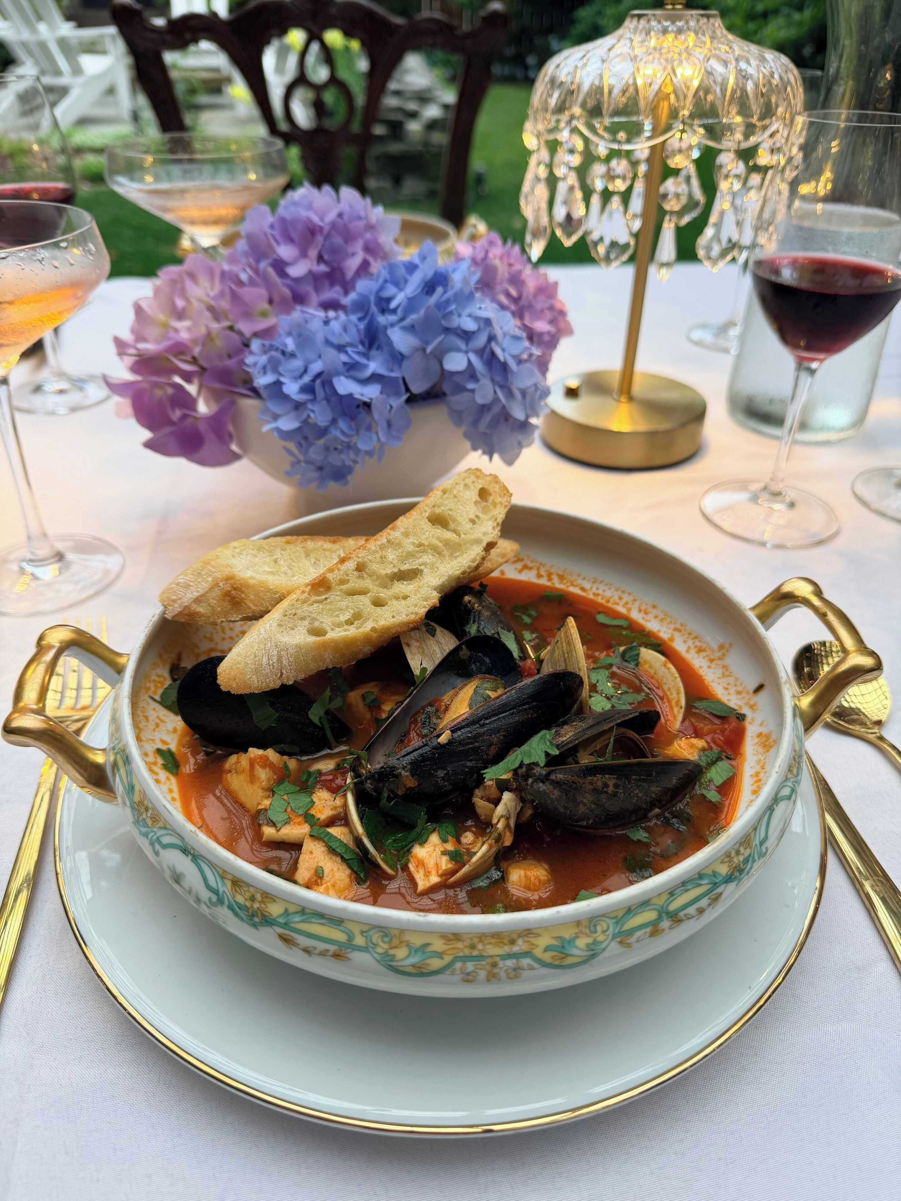 A bowl of seafood stew with bread on the side, placed on a table with a decorative vase of flowers and a candle holder.
