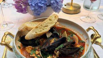A bowl of seafood stew with bread on the side, placed on a table with a decorative vase of flowers and a candle holder.