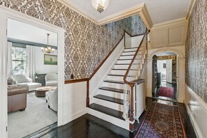 A staircase with a wooden handrail and black steps, leading to a room with patterned wallpaper and a chandelier.