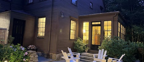 A two-story house with a stone staircase leading to a front door, surrounded by a garden with potted plants and a seating area with chairs.