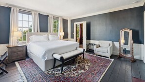 Une chambre à coucher avec un grand lit, une table de chevet, une chaise, un miroir et une fenêtre avec des rideaux.