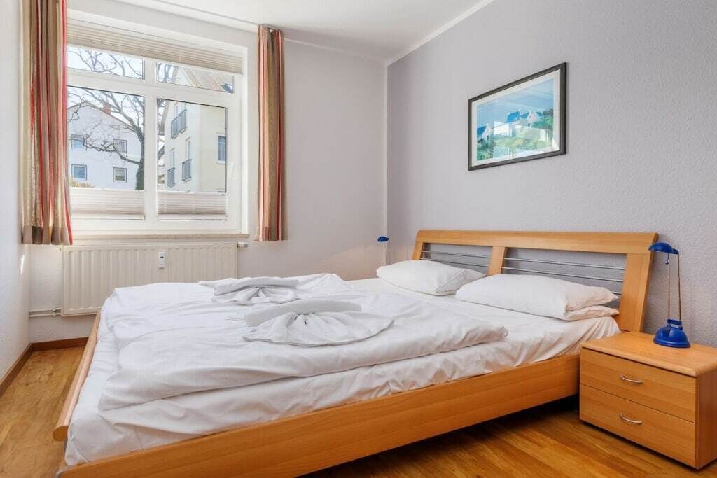 A neatly made bed with white linens, a wooden headboard, a bedside table with a lamp, and a framed picture on the wall.