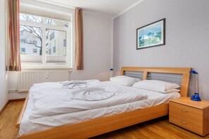 A neatly made bed with white linens, a wooden headboard, a bedside table with a lamp, and a framed picture on the wall. - 03, Residence Dünenstraße (Binz)
