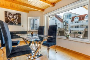 A modern dining area with a glass table and black chairs, large windows with a view of buildings, and a wooden ceiling. - Holiday apartment in Binz with swimming pool (Binz)