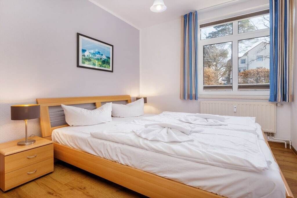A neatly made bed in a room with a single nightstand, a framed picture on the wall, and a window with curtains.
