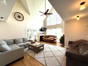 A spacious living room with a high ceiling, a fireplace, a television, and a large window.